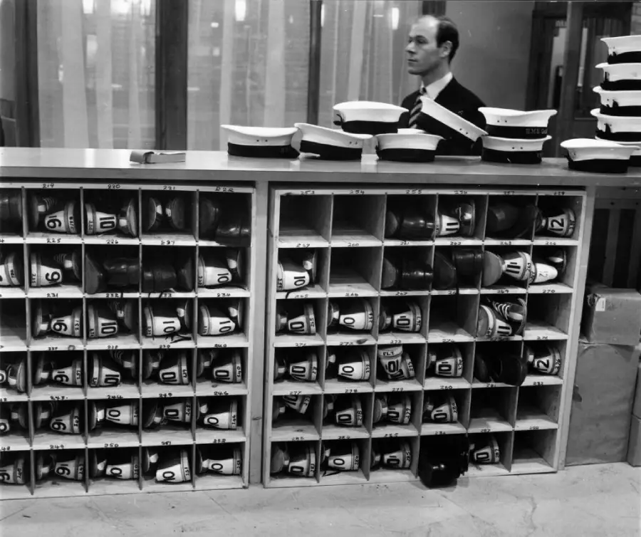 A man stands behind a counter with numbered cubbyholes holding hats and other items. More hats are stacked on top of the counter and cubbies. The background shows glass panels and curtains.