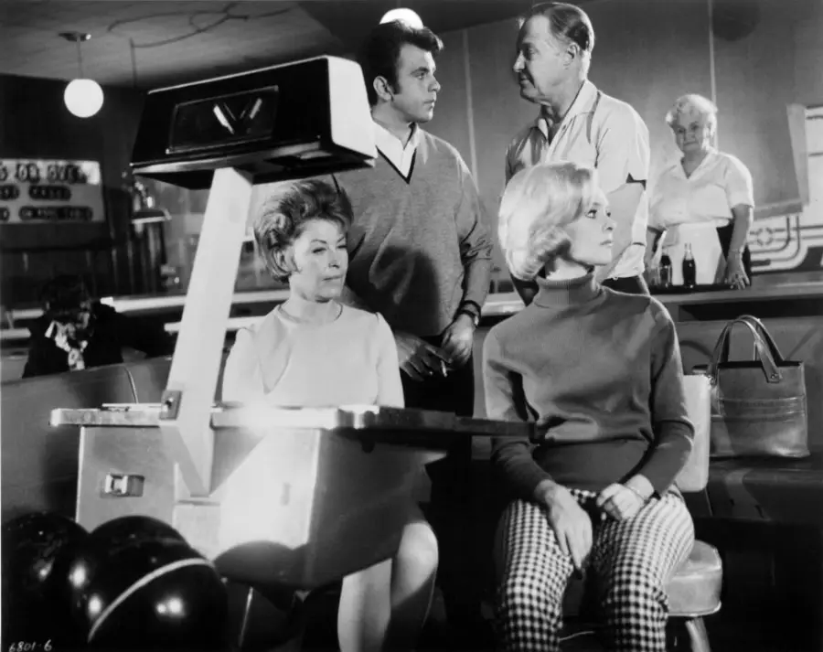 Two women sit at a bowling alley, one in a light dress and one in a sweater and checkered pants. A man stands behind them, talking to another man, while an older woman stands in the background near bowling lanes.