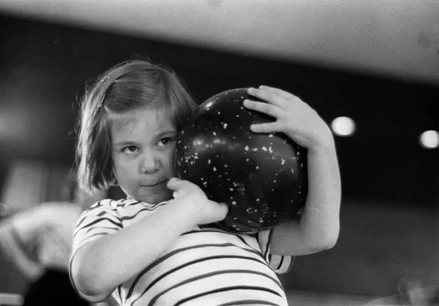 A young girl in a striped shirt holds a large bowling ball close to her face, concentrating as she prepares to bowl.