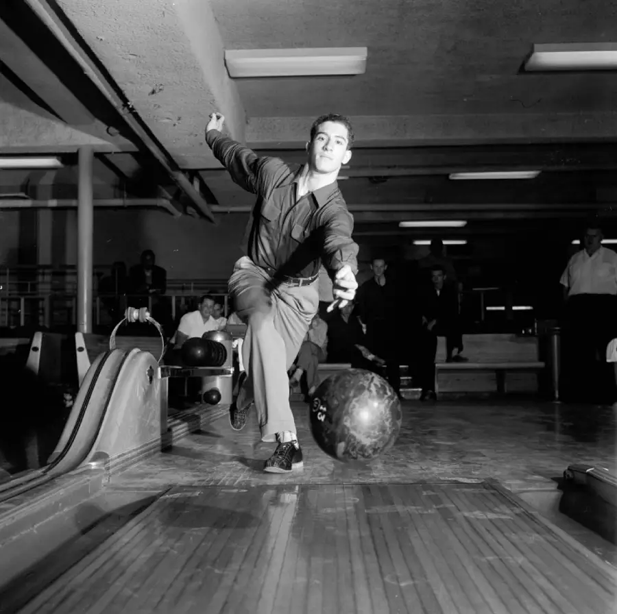 A man in mid-motion bowls a ball down a lane in a bowling alley. He wears a dark outfit and is focused, with people and benches visible in the background.