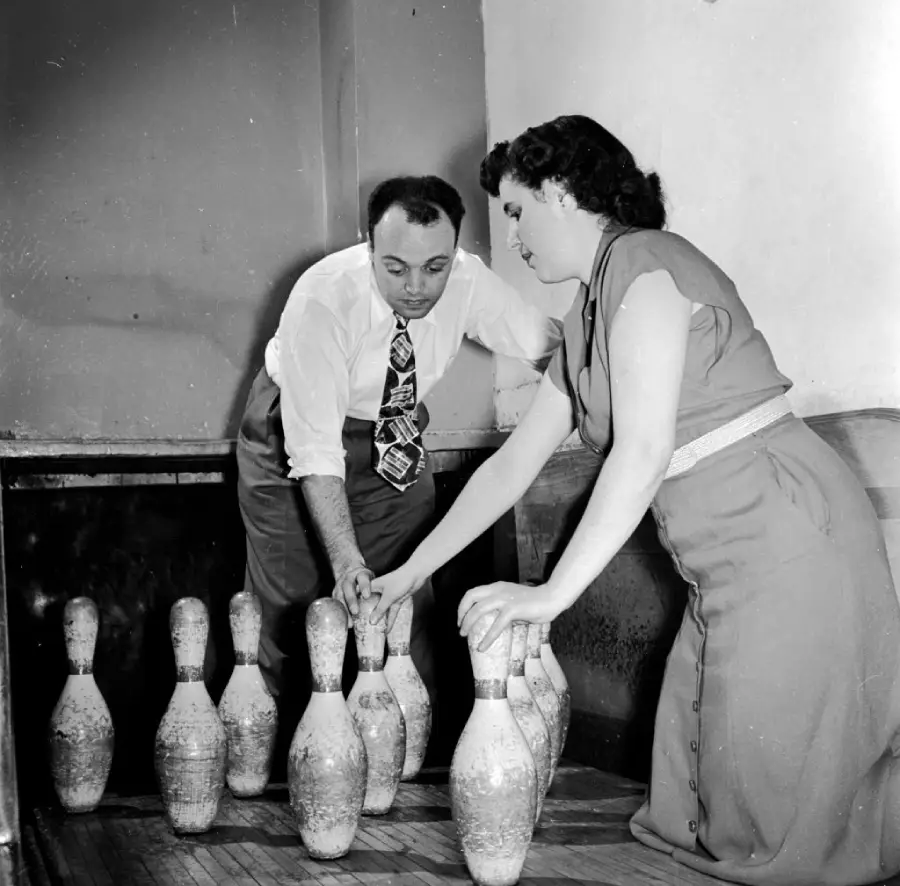 A man and a woman are setting up bowling pins by hand on a wooden lane. Both are focused on placing the pins in position. The scene appears to be from a past era, judging by their vintage clothing.