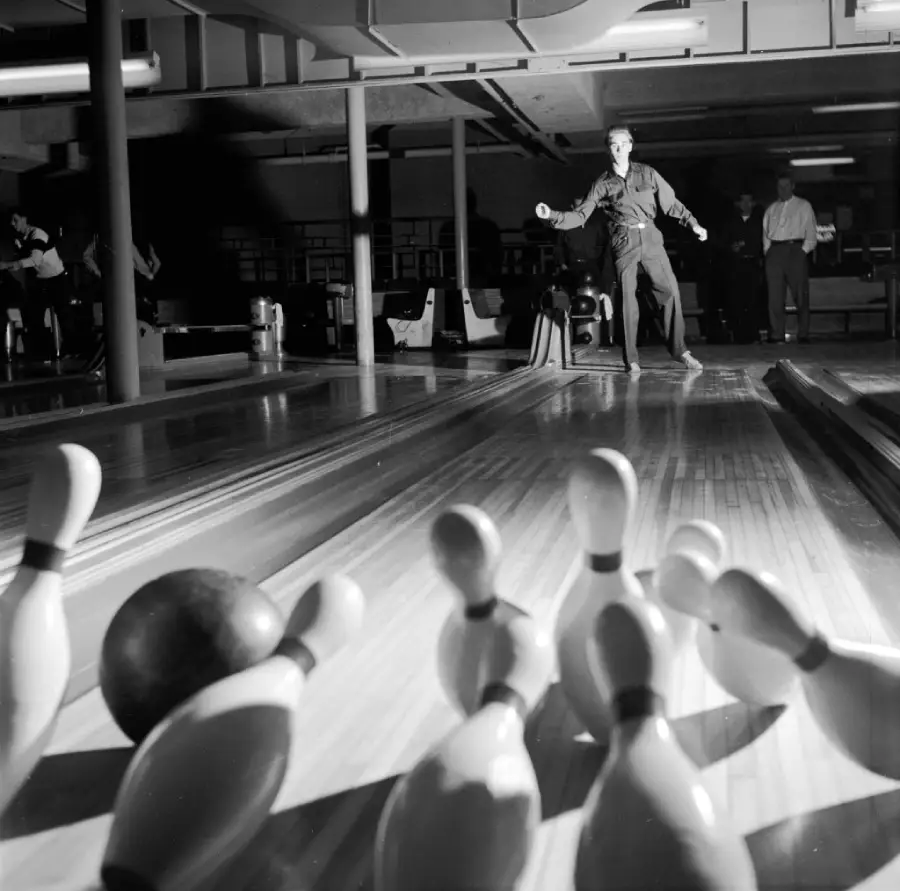 A person bowls in a dimly lit alley as pins scatter from a black bowling ball’s impact. Several people watch in the background.