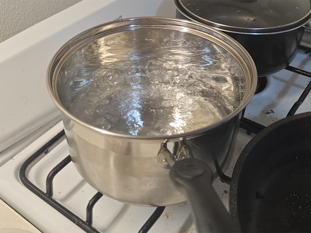 A stainless steel pot filled with boiling water sits on a gas stove, with bubbles rising to the surface and another pot and pan nearby.
