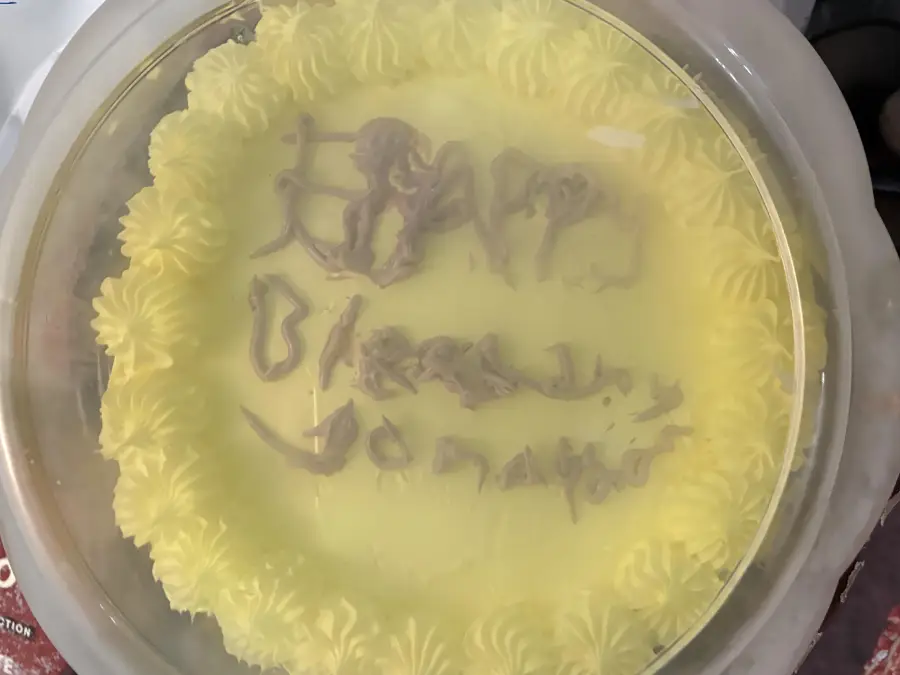 A round yellow cake with unclear, messy writing on top, covered by a plastic lid. The cake has piped icing along the edge and the text is difficult to read.