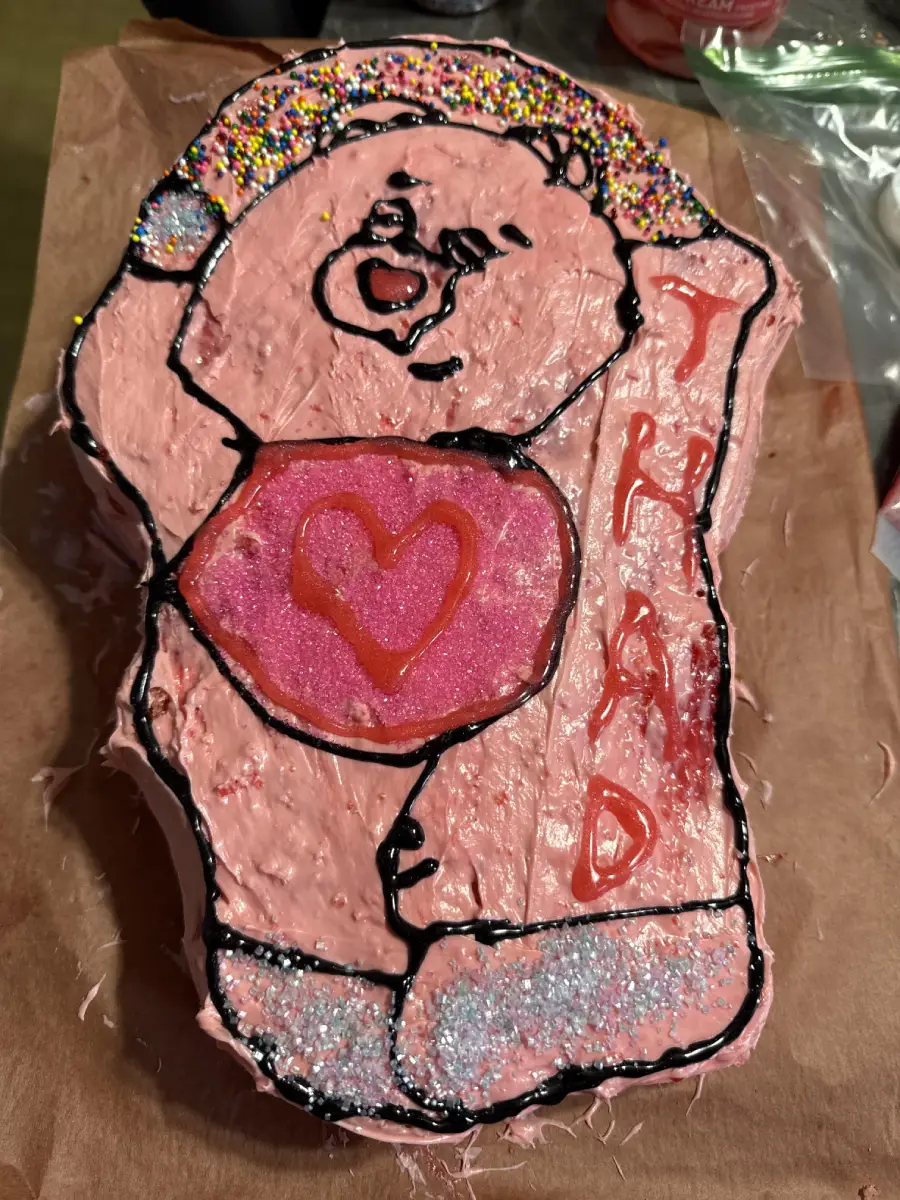 A bear-shaped cake with pink icing, sprinkles on the ear, and a heart on its belly. The word “THAD” is written in red icing down the side. The cake sits on brown parchment paper.