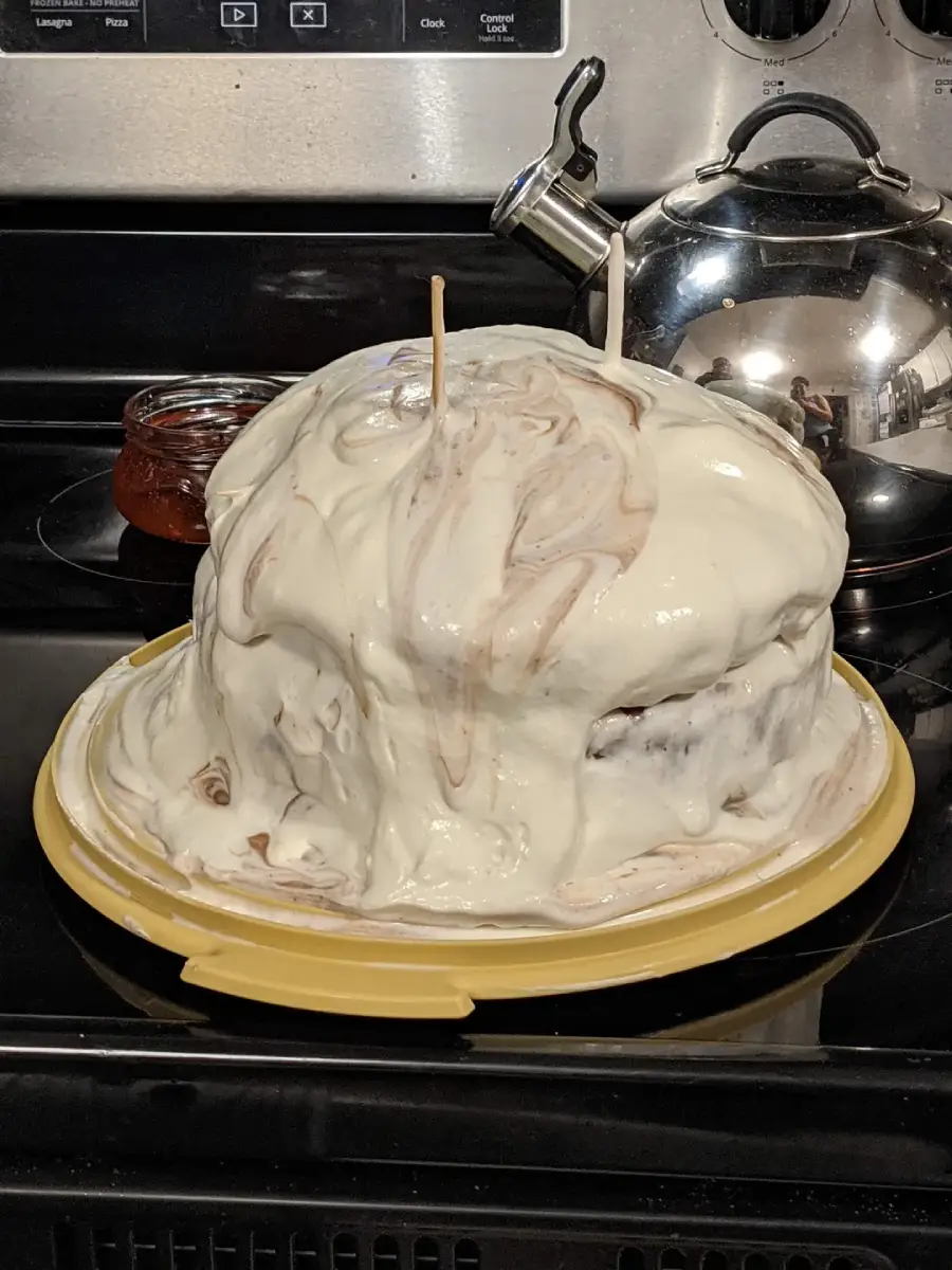 A homemade cake with uneven white and brown marbled icing sits on a yellow tray. Two wooden sticks protrude from the top. The cake is placed on a stovetop near a kettle and a jar.