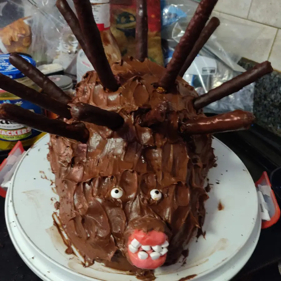 A cake shaped like a porcupine, covered in chocolate frosting with candy eyes, a red candy nose, and biscuit sticks for quills, sits on a white plate.