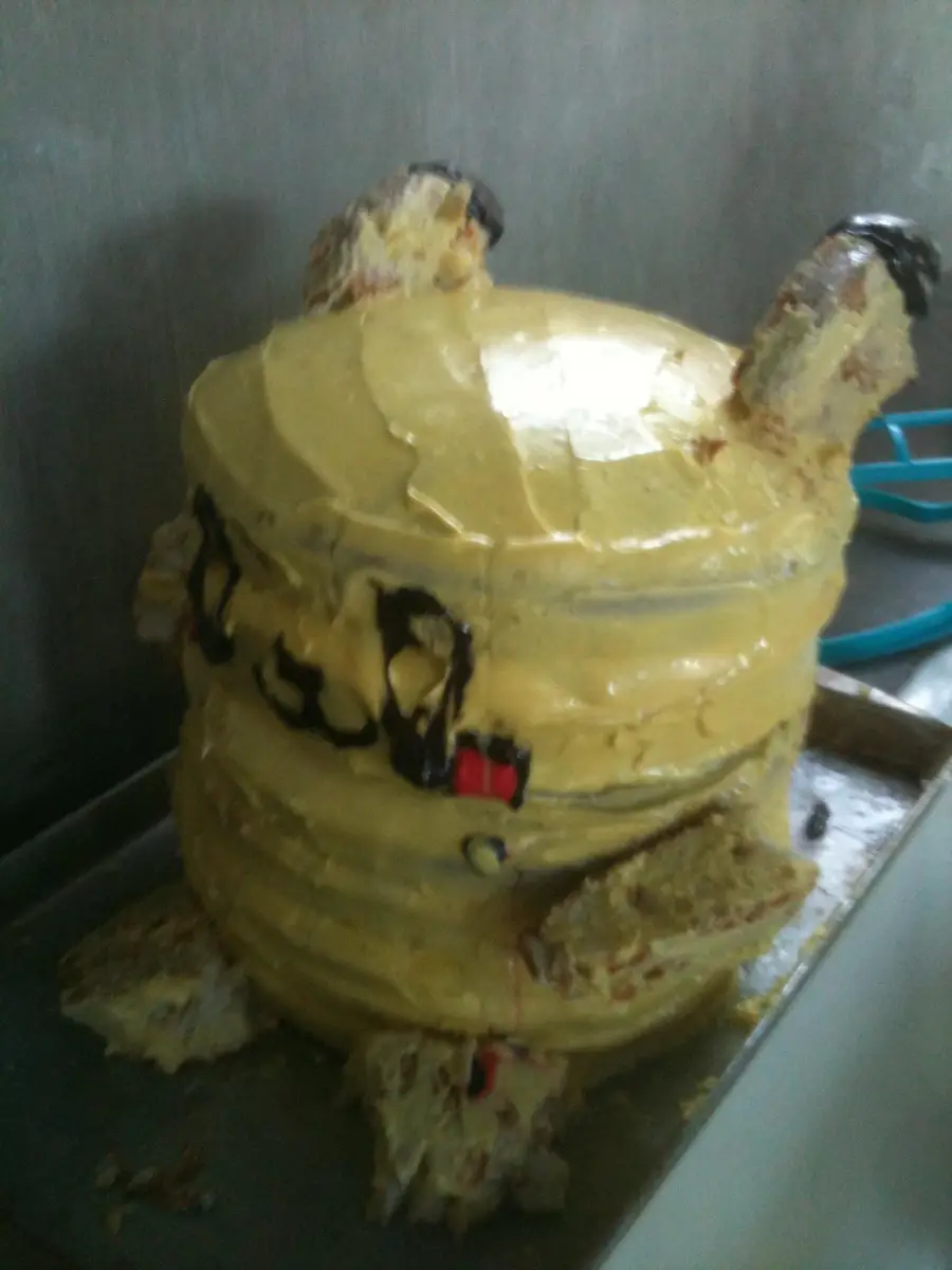 A round, yellow frosted cake with two upright ears and stubby feet, resembling a poorly made Pikachu with uneven icing and visible cake crumbs on a metal tray.