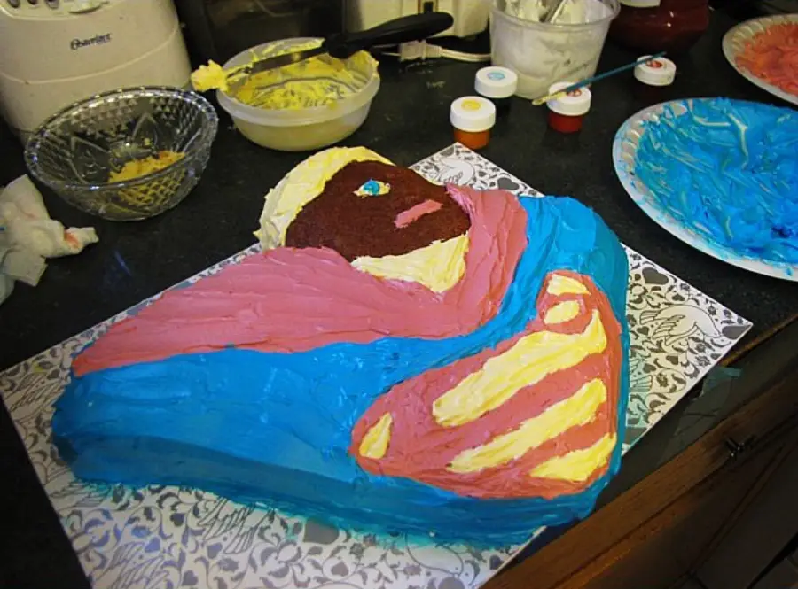 A cake decorated with colorful icing to depict a stylized person in a blue and pink outfit, lying on a patterned surface in a kitchen setting with baking supplies visible nearby.