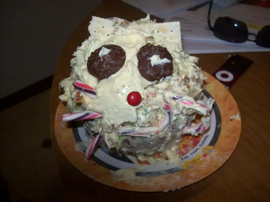 A homemade cake decorated to look like an animal face, with chocolate cookies for eyes, a red candy nose, candy sticks for whiskers, and messy white frosting on a plate.