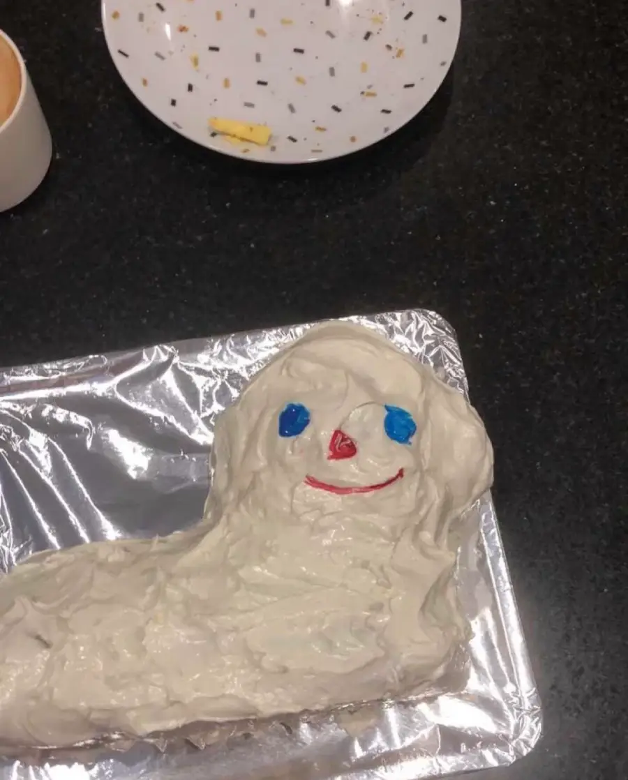 A cake shaped like a ghost or blob with white frosting, blue eyes, a red nose, and a red smile sits on foil. Behind it is a plate with sprinkles and a small piece of butter.