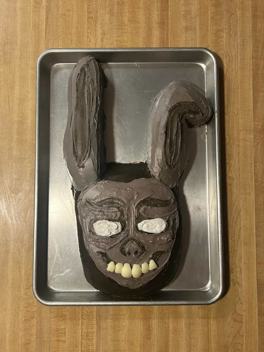A cake shaped like a creepy rabbit face with tall ears, dark frosting, white eyes, and large yellow teeth, displayed on a metal baking tray on a wooden surface.