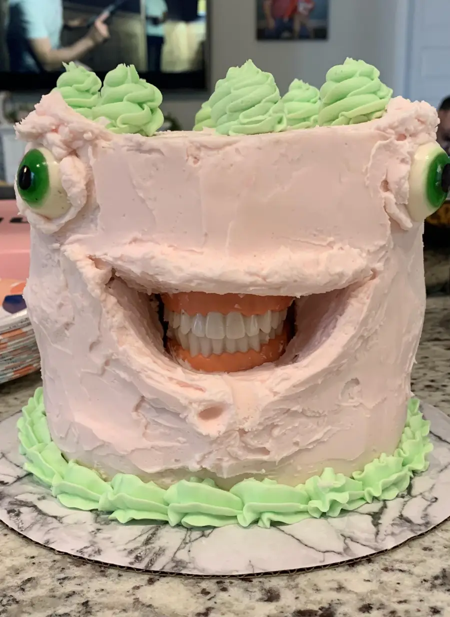 A cake decorated with pink frosting, green eyes, and green icing on top. The cake has a wide, open mouth displaying a set of realistic fake teeth, giving it a humorous and slightly unsettling appearance.