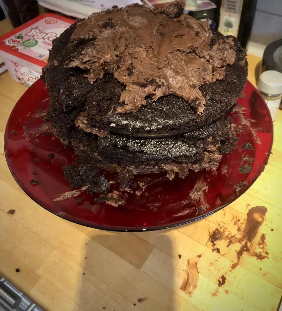 A messy, uneven chocolate cake with thick chocolate frosting, sitting on a red plate. Cake crumbs and smears of chocolate are on the plate and wooden counter.