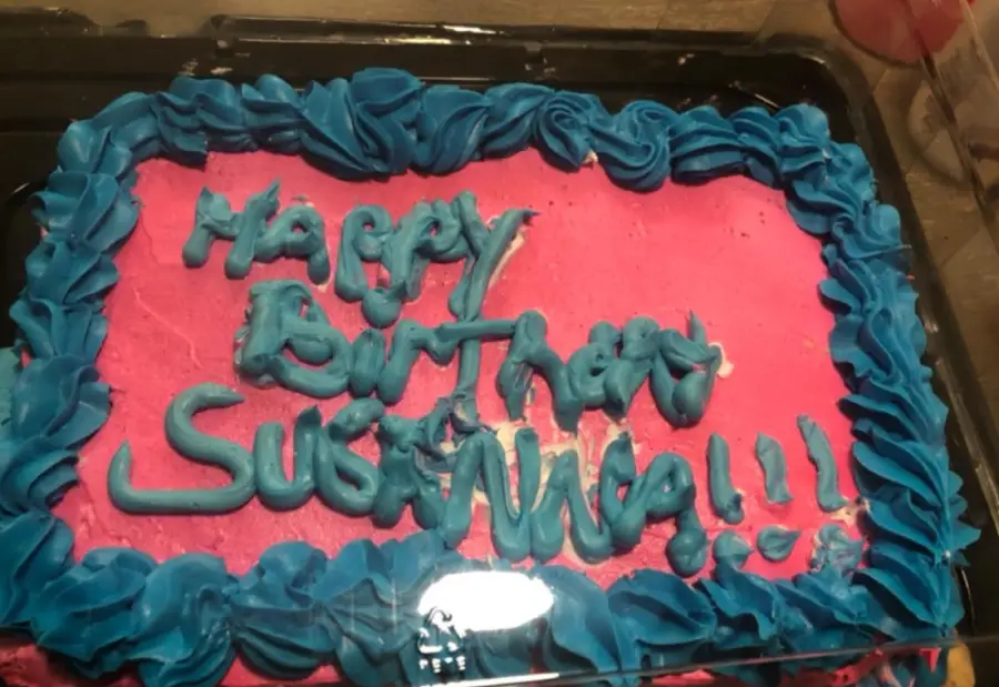 A rectangular cake with pink icing, blue icing border, and the words “Happy Birthday Susanna!!!” written in blue frosting.