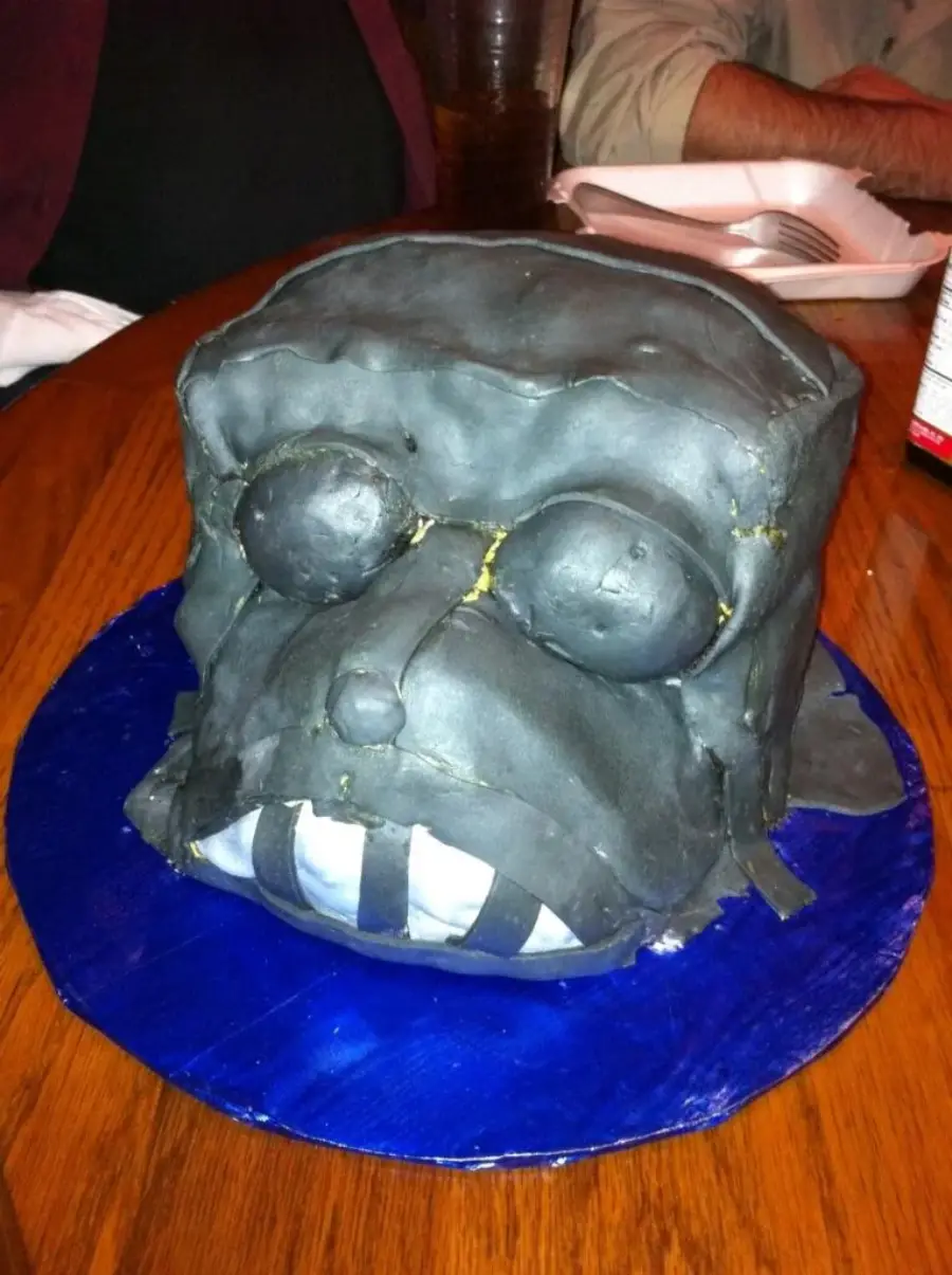 A homemade cake covered in dark gray fondant, shaped like a blocky face with bulging eyes, a protruding nose, and white teeth, placed on a blue plate on a wooden table.