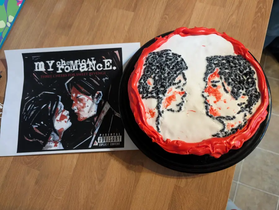 A cake decorated to resemble the “Three Cheers for Sweet Revenge” My Chemical Romance album cover sits next to a printed image of the album art on a wooden table.