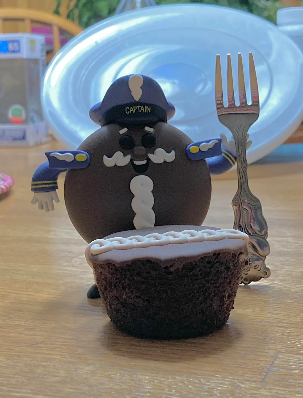 A toy captain with a round, chocolate cookie body stands behind a mini chocolate cake and a fork. A large, white plastic lid is in the background.
