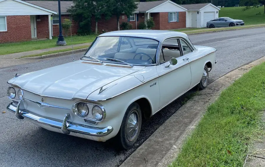 A vintage white car is parked on a residential street near a grassy curb, with brick houses and a gray car in the background.