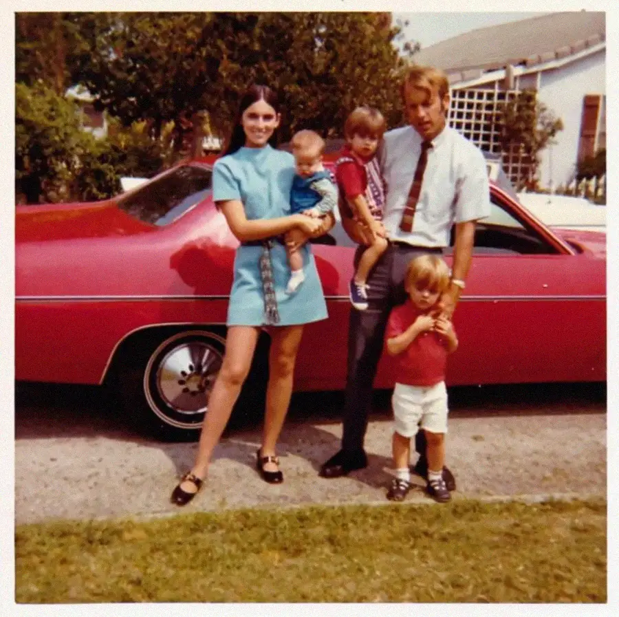 A woman and man pose in front of a red car with two young children and a baby. The woman holds the baby, and the man stands with one child while another child stands nearby. It’s a bright, sunny day in a suburban neighborhood.