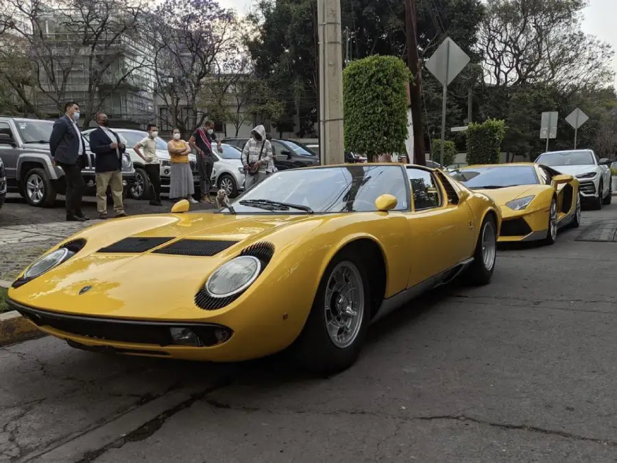 Two yellow sports cars are parked on a street, with people standing on the sidewalk nearby. Trees and other vehicles are visible in the background.