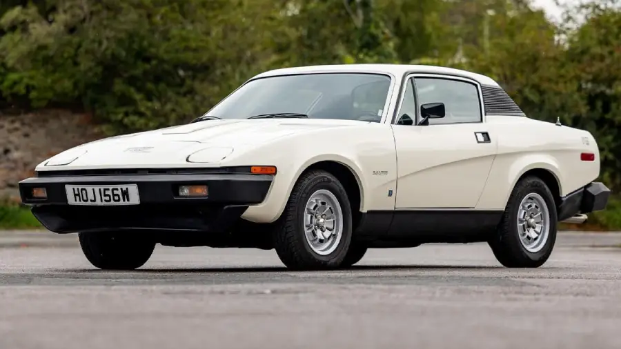 A white classic sports car parked on a paved surface, with trees and greenery in the background. The car has a sleek design, round headlights, and a black front bumper.