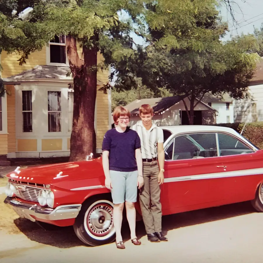 Two smiling kids stand in front of a shiny red classic car parked on a sunny residential street, with trees and a yellow house in the background.