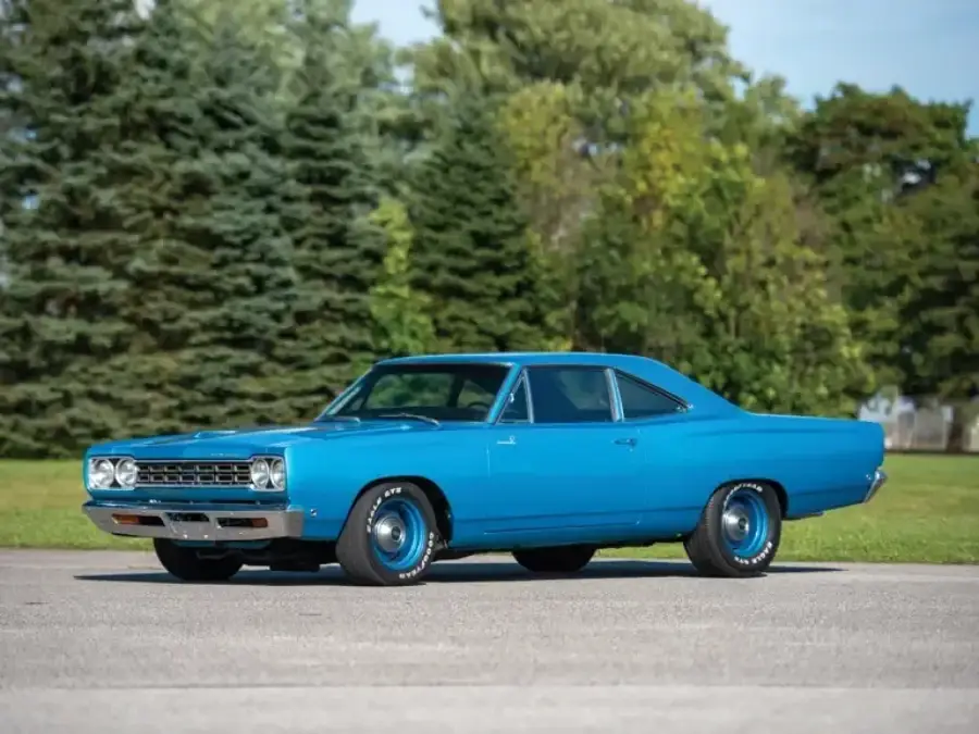 A vintage blue muscle car is parked on a paved surface with green grass and tall trees in the background under a clear sky.