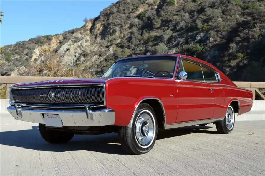 A classic red muscle car with chrome accents is parked on a sunlit road, with rocky hills and trees in the background.