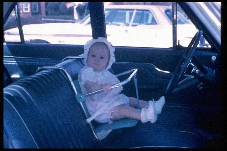 A baby in a white outfit and bonnet sits in a car’s front seat, secured in an old-fashioned metal car seat. The baby holds onto the frame, with parked cars visible outside the window.