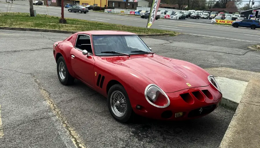 A classic red sports car with a sleek, curved body is parked in a lot near a busy street. Several cars and buildings are visible in the background.