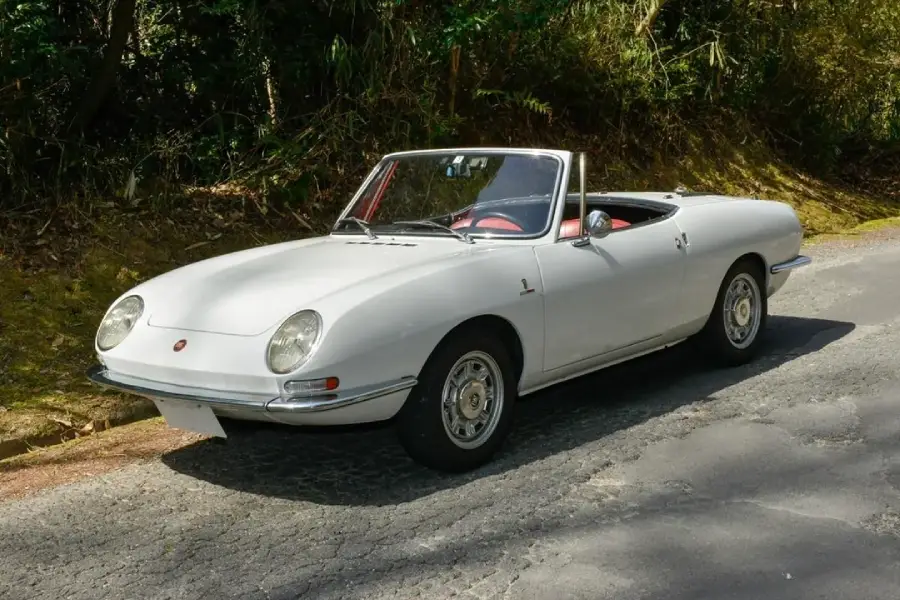 A classic white convertible car with a red interior is parked on a shaded road beside greenery. The car has round headlights and a sleek, vintage design.