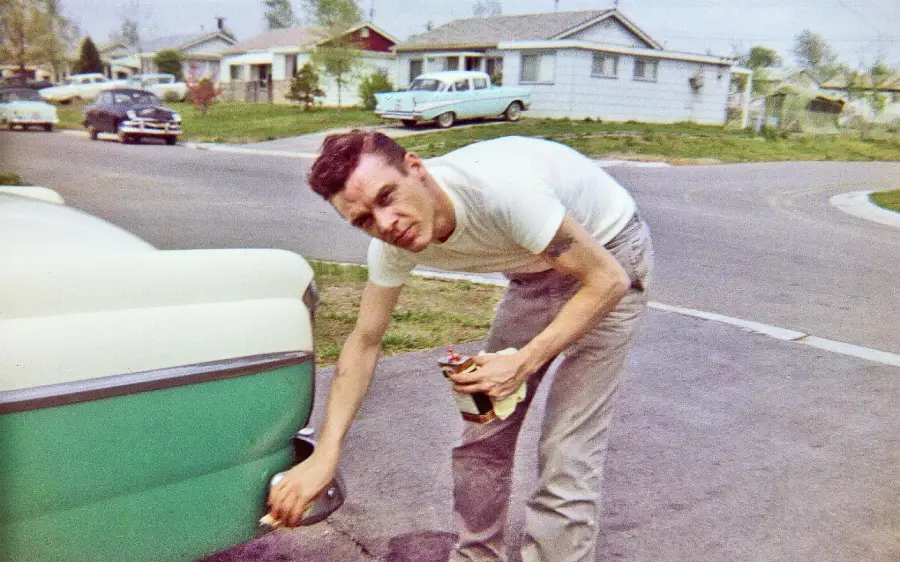 A man in a white T-shirt and gray pants is cleaning the back of a green and white car on a suburban street, holding a cloth and a bottle. Houses and vintage cars are visible in the background.