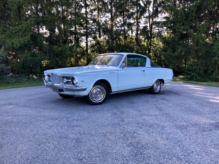 A light blue classic car is parked on a paved area with trees in the background under clear daylight.