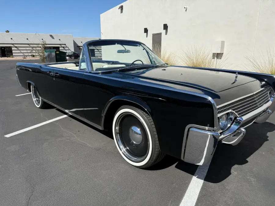 A classic black convertible car with whitewall tires is parked in a lot near a beige building under a clear blue sky.