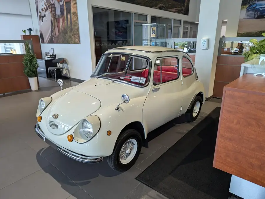 A vintage white compact car with round headlights and red interior is displayed inside a modern showroom with large windows and wood accents.
