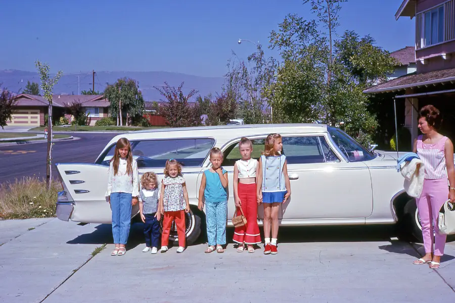 Six children and a woman stand in front of a white classic station wagon parked in a suburban driveway on a sunny day. The children are dressed in colorful 1970s clothing. The background shows houses and trees.