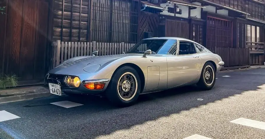 A silver classic sports car is parked on a street in front of traditional wooden buildings, with its headlights on and vintage-style wheels visible.