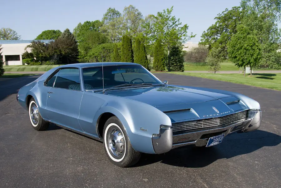 A classic blue vintage car with sleek, rounded edges is parked on a paved driveway, surrounded by green grass, trees, and a few buildings in the background under a clear sky.