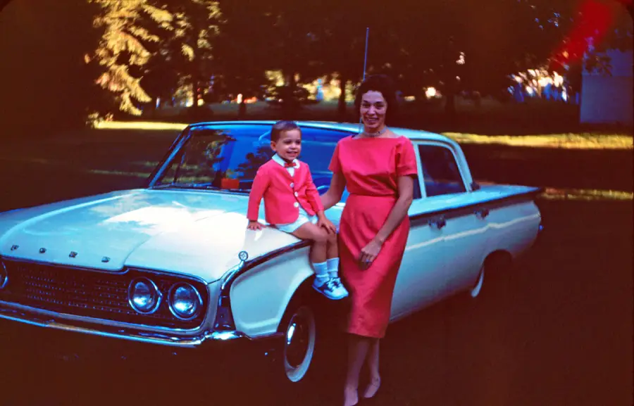A smiling woman in a red dress stands next to a young child sitting on the hood of a vintage white car. The child wears a red jacket and shorts. Trees and grass are visible in the background.