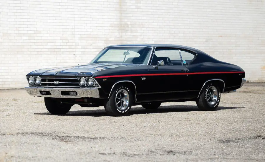 A black classic muscle car with chrome details, a red stripe along the side, and SS badges is parked on a paved surface in front of a white brick wall.