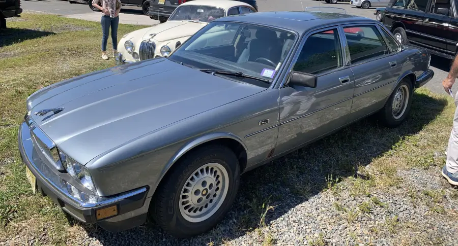 A silver classic sedan is parked on gravel, with another vintage car and a few people in the background on a sunny day.