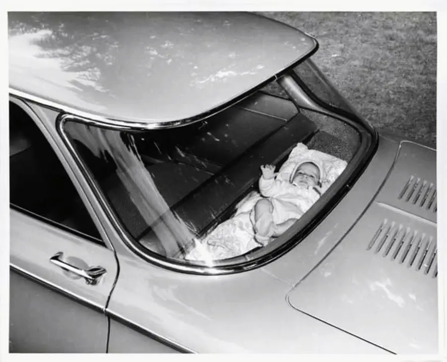 A baby lies on a blanket in the rear window shelf of a vintage car, viewed from above. The car is parked outdoors.