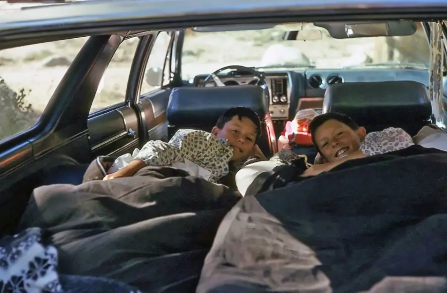Two children lie under blankets in the back of a car, smiling at the camera. The car seats have been folded down to make a cozy sleeping area, and daylight streams in through the windows.