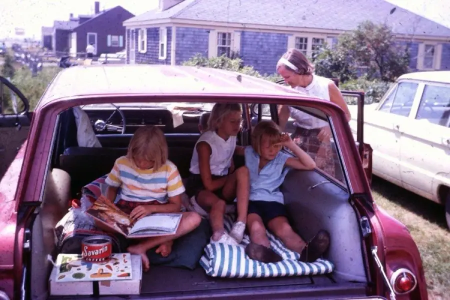 Three children sit in the back of an open red station wagon, reading and relaxing. A woman stands nearby outside. The scene appears to be from the past, with vintage cars and houses in the background on a sunny day.