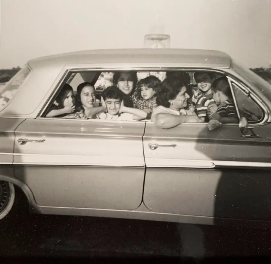 A black and white photo shows eight children crowded inside an old car, smiling and looking out the windows on a sunny day.