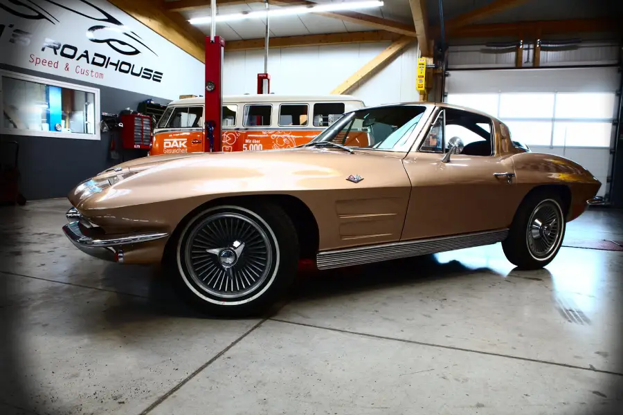 A gold classic Corvette Stingray coupe is parked indoors next to an orange and white vintage Volkswagen bus under exposed wooden beams.