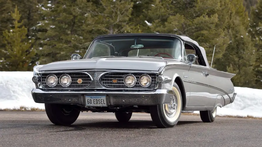 A classic silver 1960 Edsel convertible car is parked on a road, with snow on the ground and green trees in the background. The license plate reads “60 EDSEL.”