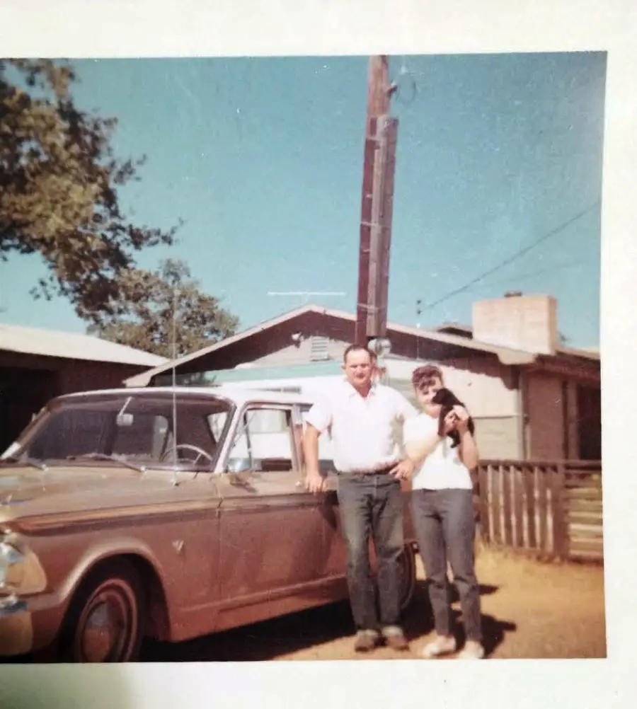 A man and woman stand next to a vintage beige car outside a house on a sunny day. Trees and a wooden fence are visible in the background.