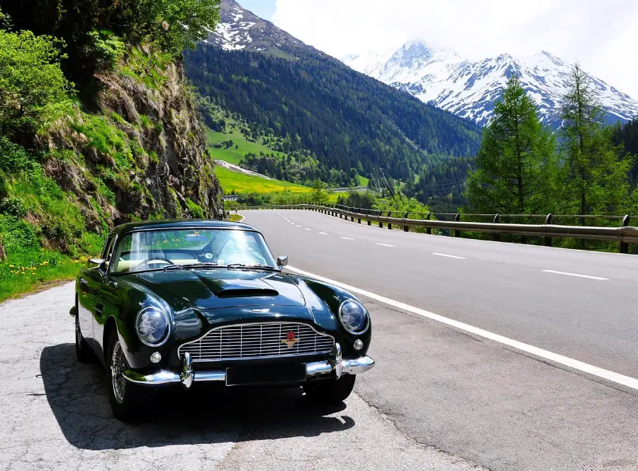 A classic dark green convertible car is parked on a mountain road, with snowy peaks, lush green trees, and a clear sky in the background.